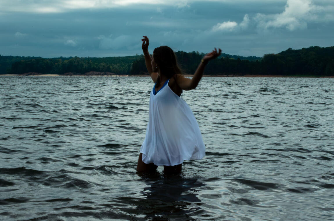 A figure in a white dress standing waist-deep in a lake with both arms raised under a storm sky